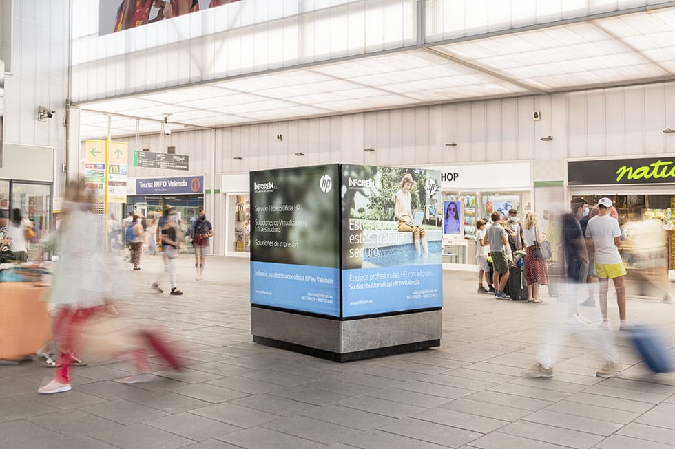 Publicidad de Inforein en la estación AVE de Valencia con pasajeros y tiendas al fondo.